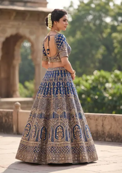 A beautiful Indian bride viewed from behind, wearing a royal blue lehenga heavily embroidered with gold patterns and a backless blouse with tassels, standing outside an old stone palace.