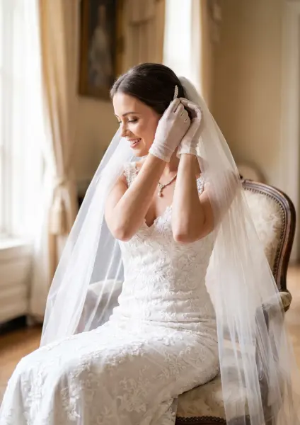 A happy bride wearing a lace wedding dress, a long veil, and white satin gloves, sitting in an ornate vintage chair and smiling while adjusting her hair.
