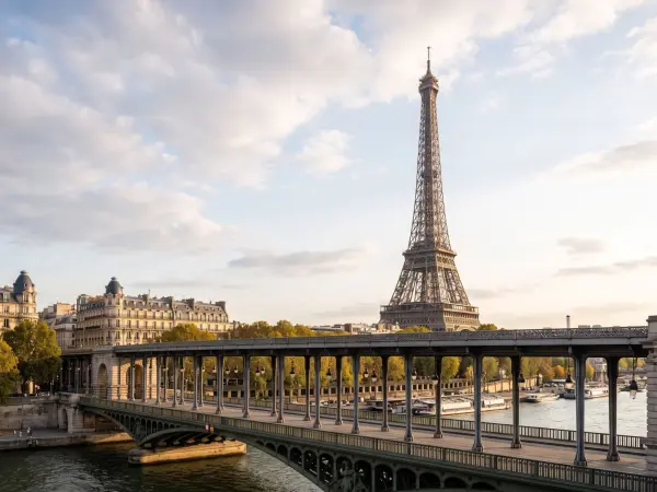 A wide shot of the iconic Eiffel Tower towering over the Seine River, viewed from an angle that prominently features the ornate steel columns of the Pont de Bir-Hakeim bridge in the foreground under a partly cloudy sky.