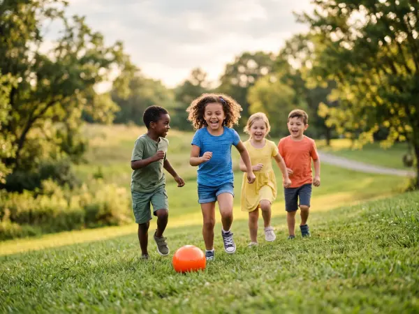 Four young, diverse children running and smiling enthusiastically towards the camera across a green grassy park area on a sunny day, chasing a bright orange soccer ball.