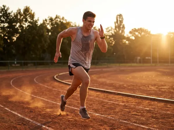 A sweating, determined male runner sprinting hard on an outdoor red running track, with the bright golden light of the setting sun shining in the background.