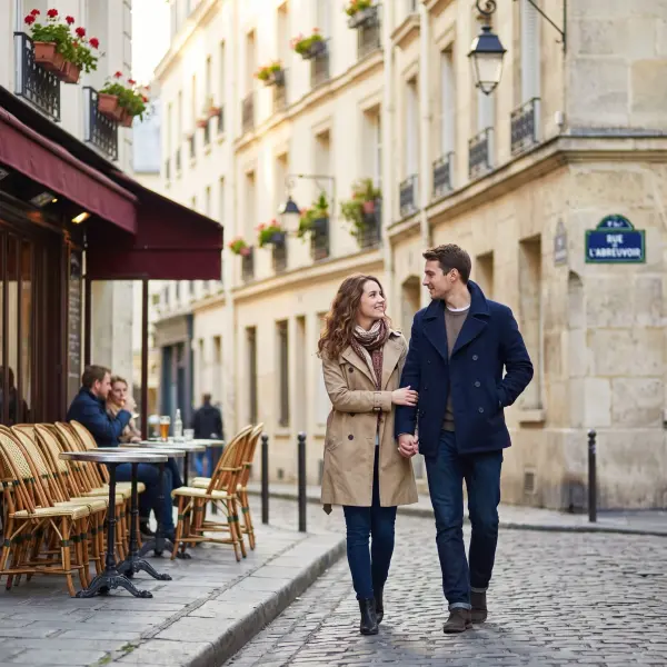 A stylishly dressed man and woman holding hands and walking towards the camera on a narrow cobblestone street in Paris, with traditional bakeries and cafes in the background.