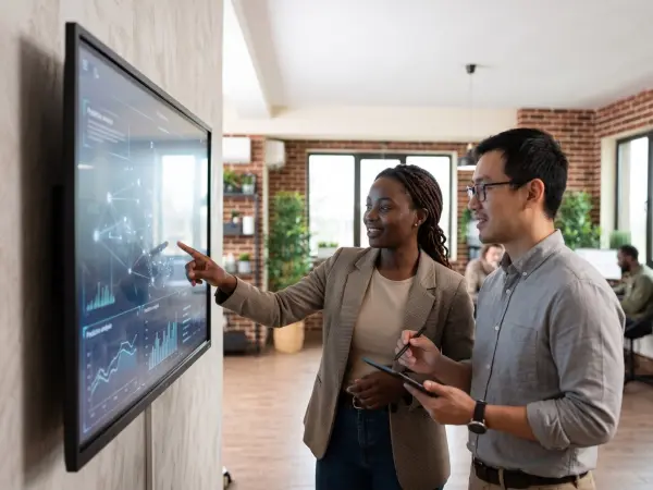 A Black businesswoman and an Asian businessman standing in a modern office, smiling and collaborating while pointing at complex data charts displayed on a large interactive digital smart board mounted on the wall.